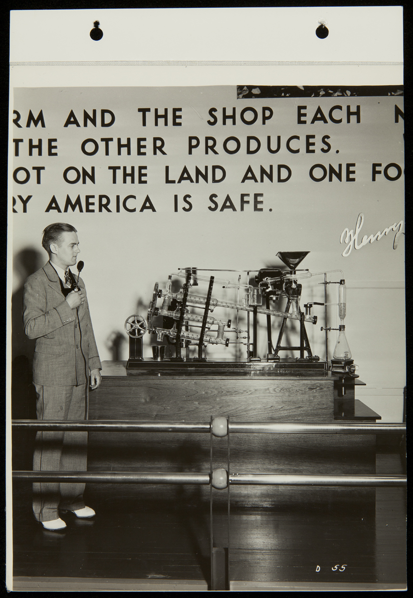 Person in suit holding microphone stands next to a piece of equipment under text on a wall