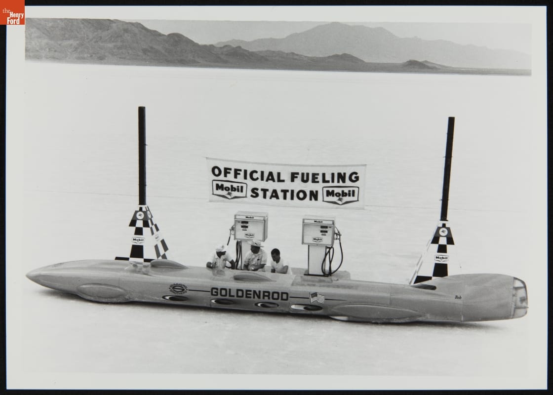 Goldenrod Land Speed Race Car, circa 1965 Long, low, race car at gas pumps under banner reading "Official Fueling Station," in open area with mountains in the background