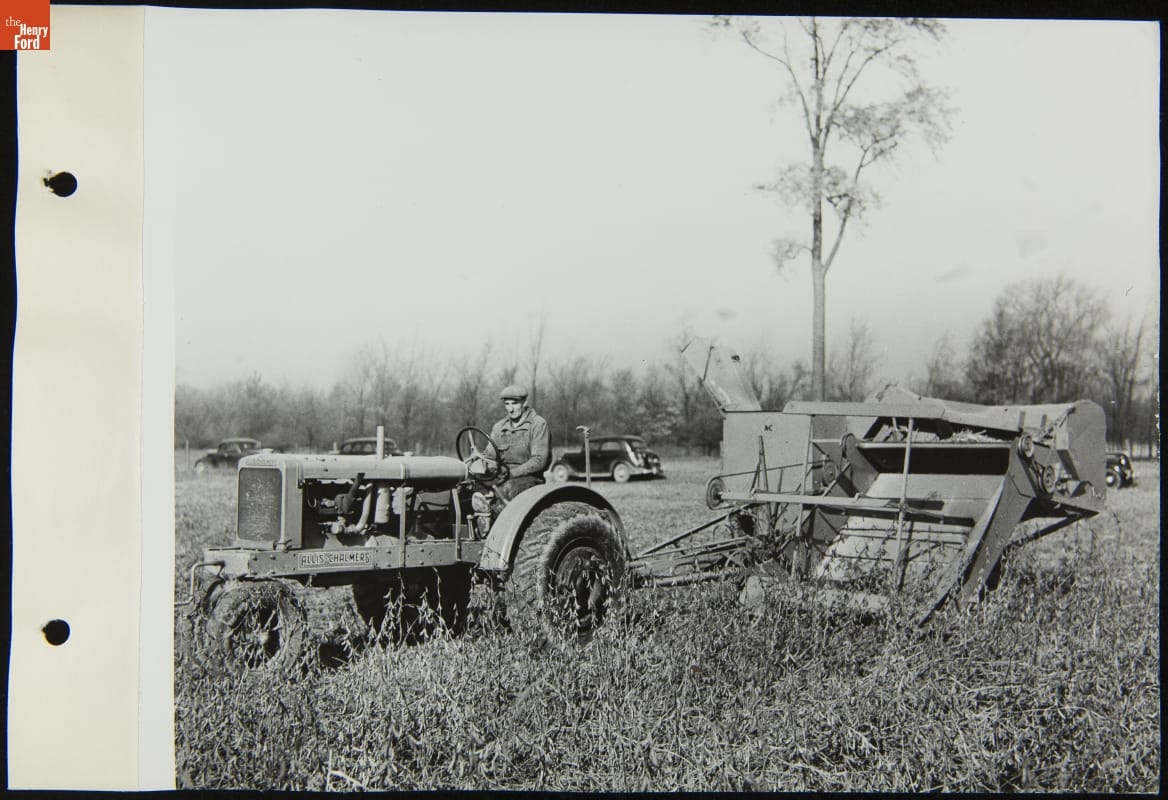 Black-and-white photo of man driving a tractor with large equipment attachment behind it through a field