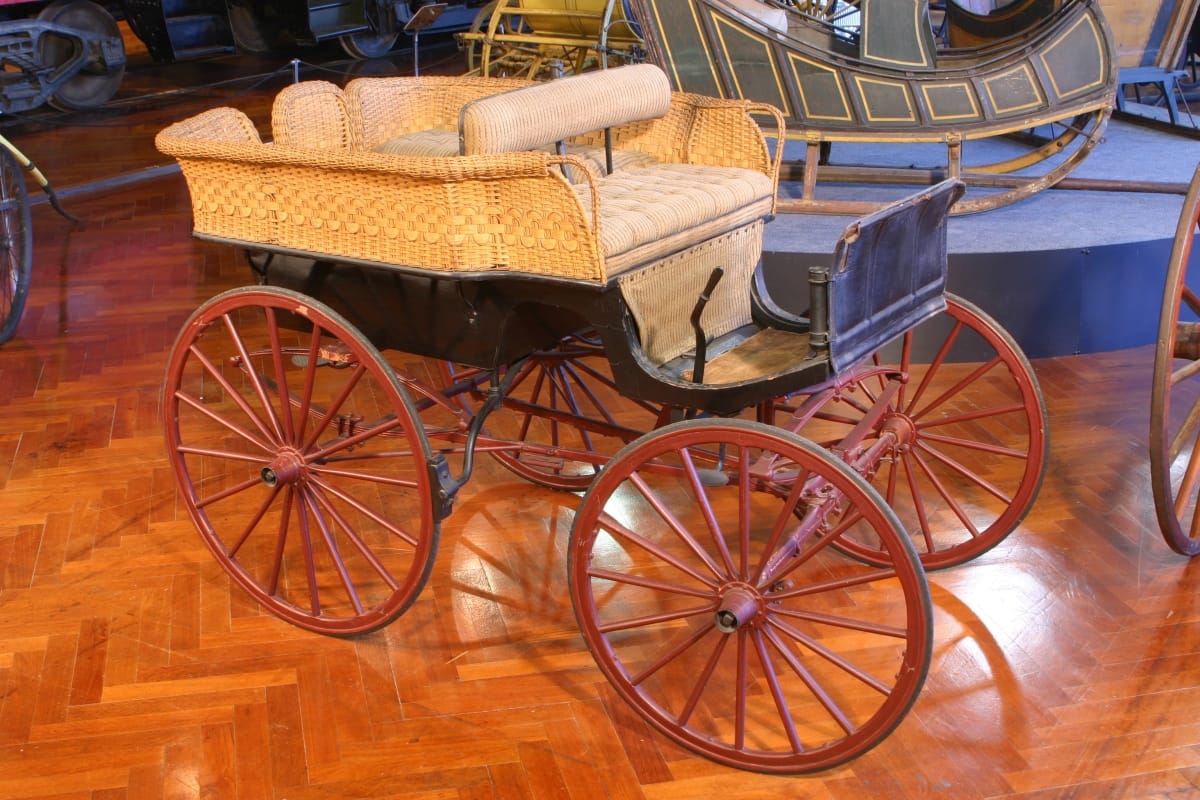 Black open carriage with red wheels and beige rattan (?) sides, displayed in a room with other vehicles