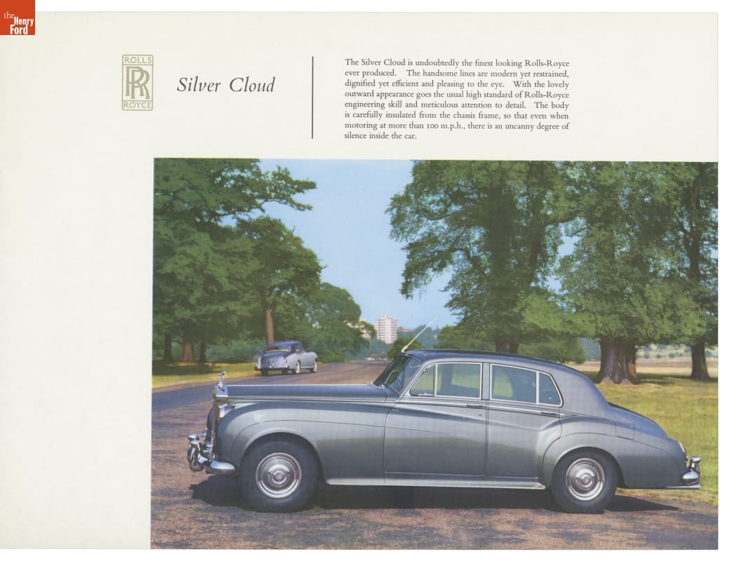 Silver limousine with long hood parked crossways on driveway with grass, trees, buildings in the vicinity