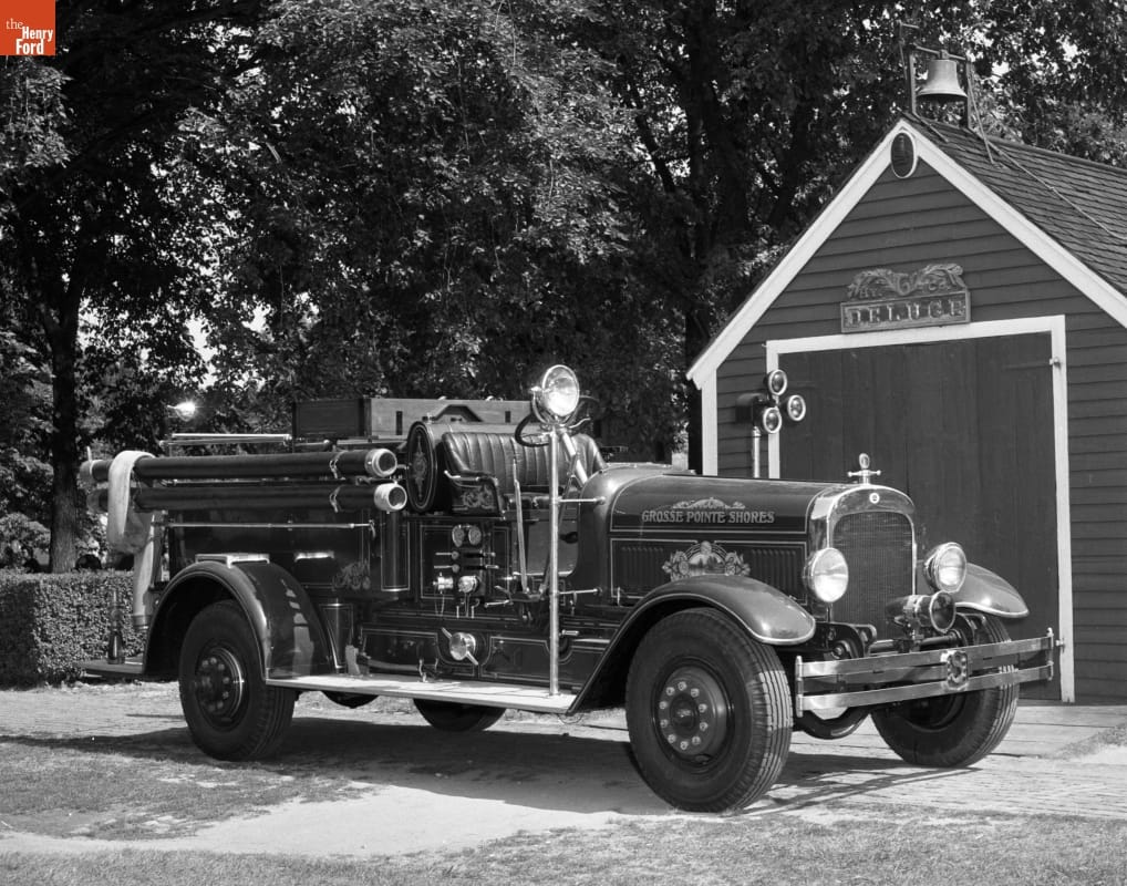 Black-and-white photo of fire engine parked in front of wooden shed or garage and trees