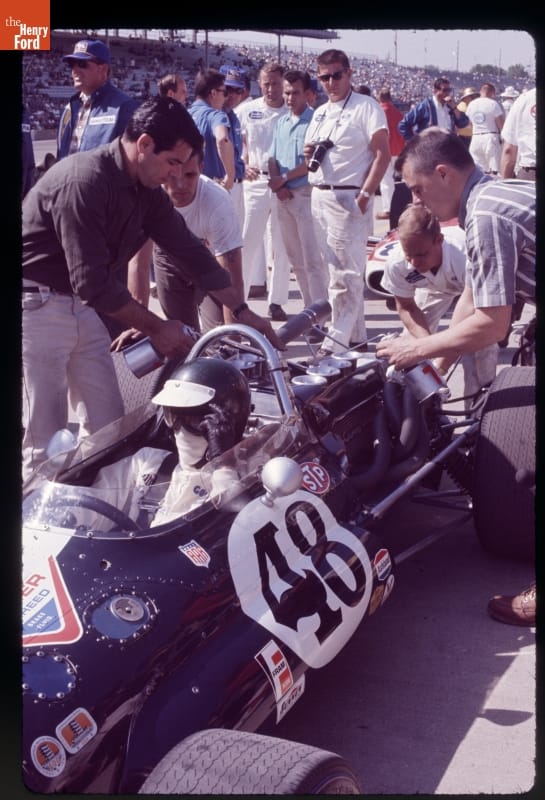 Man sitting in race car with several people gathered around it; additional people in the middle distance and grandstands full of people in the far background