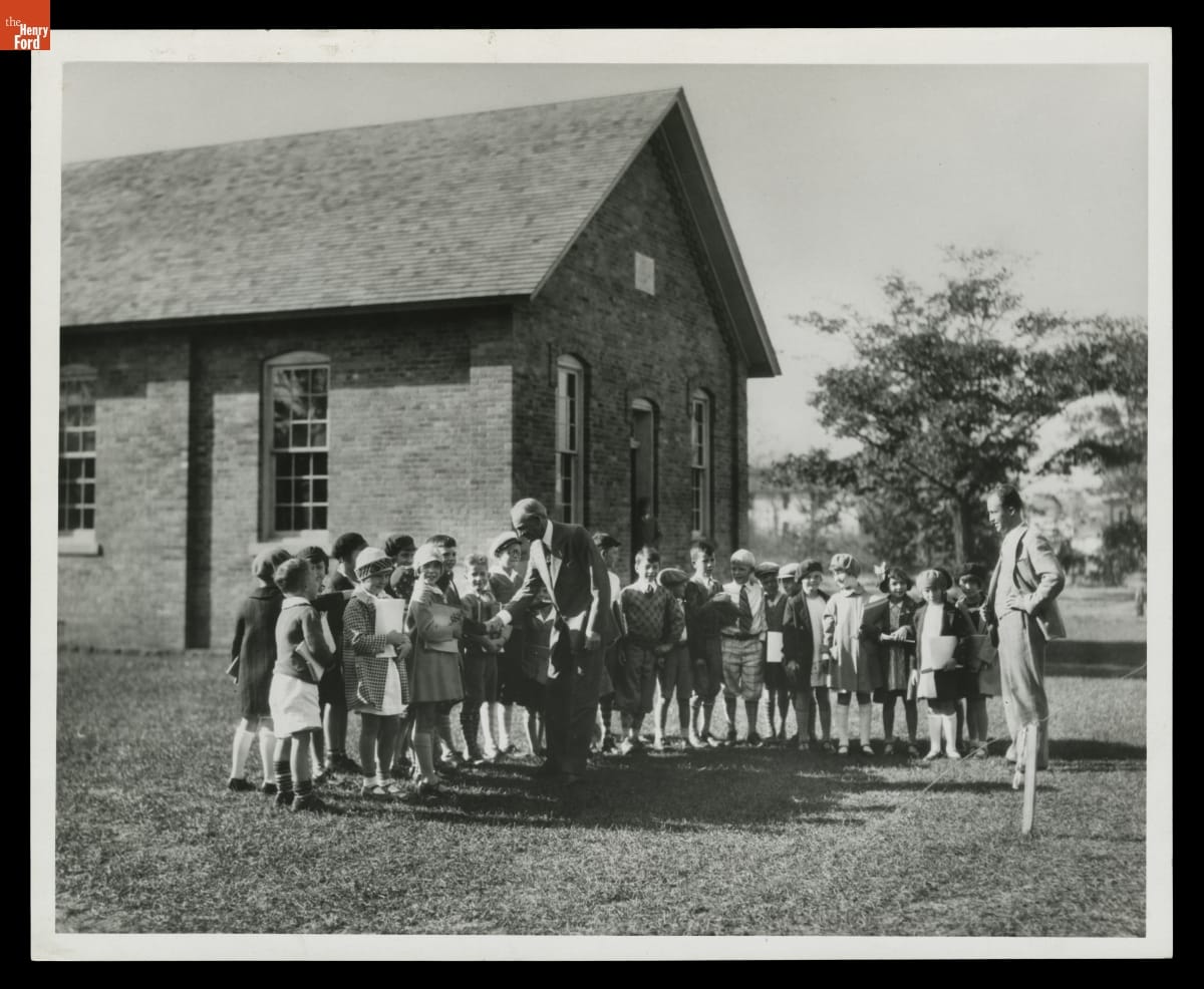Henry Ford with Students outside Scotch Settlement School in Greenfield Village, 1929 Two men stand with a group of children outside a small brick building; one shakes hands with a child