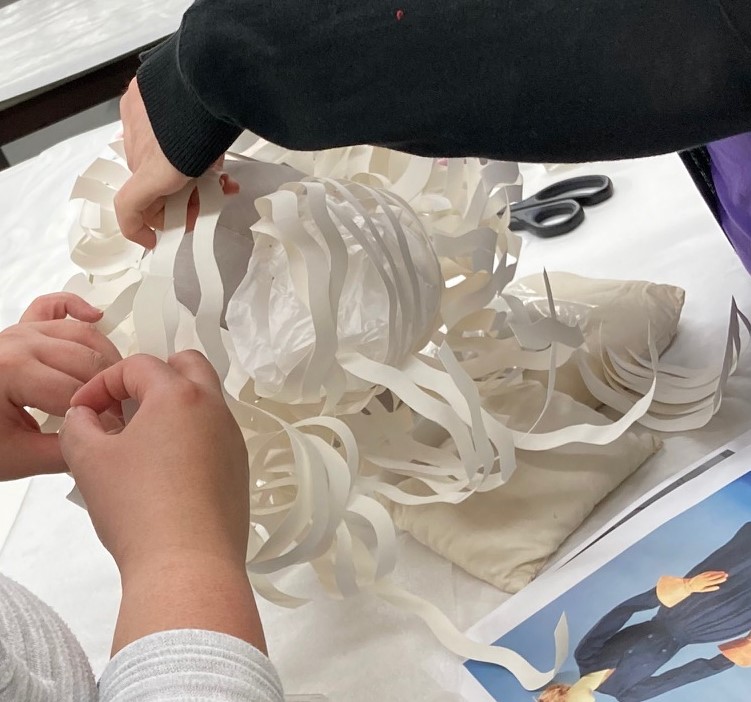 An arm extends, holding up a complex white cut paper shape, while two hands in lower left also hold the shape