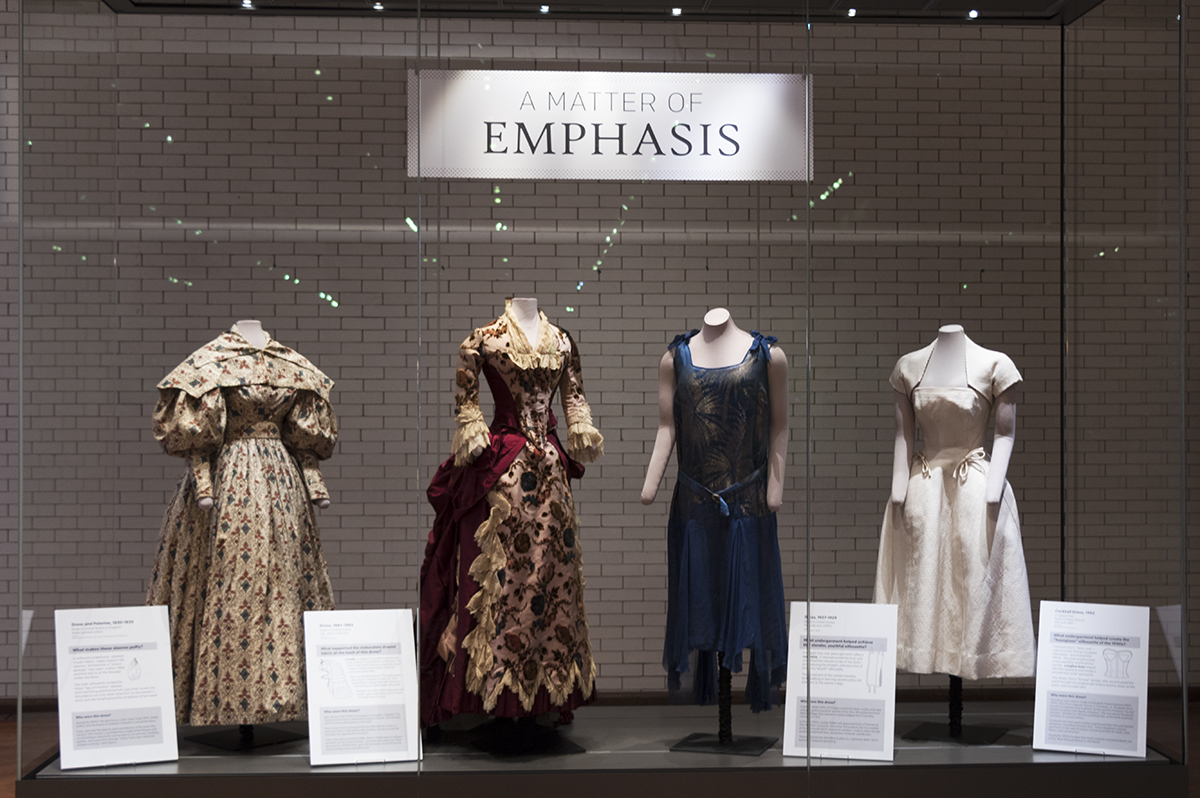 What We Wore: A Matter of Emphasis exhibit case in Henry Ford Museum of American Innovation Display case containing four dresses on mannequins, each with a sign nearby